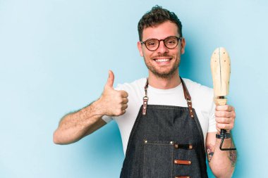 Kafkasyalı genç kunduracı mavi arka planda izole edilmiş gülümsüyor ve başparmağını kaldırıyor.