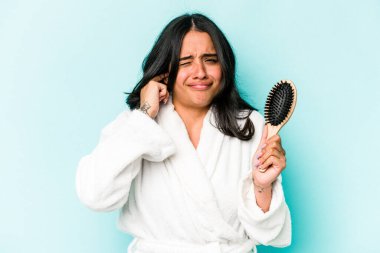 Mavi arka planda saç fırçası tutan genç İspanyol kadın kulaklarını elleriyle kapatıyor..