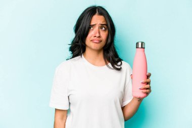 Mavi arka planda termosu olan genç İspanyol kadın şaşkın ve kararsız hissediyor..