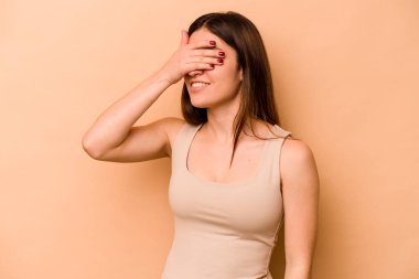 Latin kökenli genç bir kadın, bej arka planda izole edilmiş. Elleriyle gözlerini kapatıyor. Gülümsüyor ve bir sürpriz bekliyor..