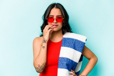 Elinde plaj havlusu tutan genç İspanyol kadın mavi arka planda izole edilmiş tırnak yiyor, gergin ve çok endişeli..