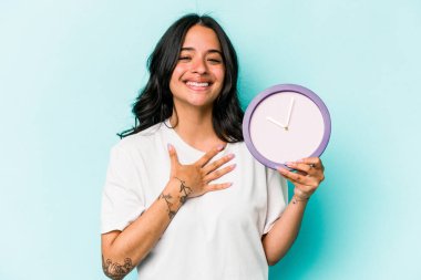 Mavi arka planda izole edilmiş bir saati tutan genç İspanyol kadın elini göğsünde tutarak kahkahalar atıyor..