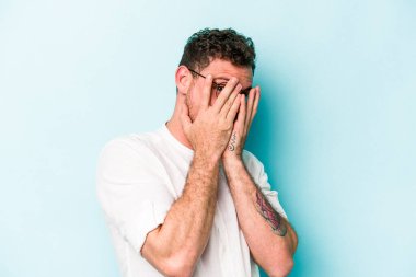 Kafkasyalı genç adam mavi arka planda izole edilmiş. Korkmuş ve gergin parmaklarıyla göz kırp..