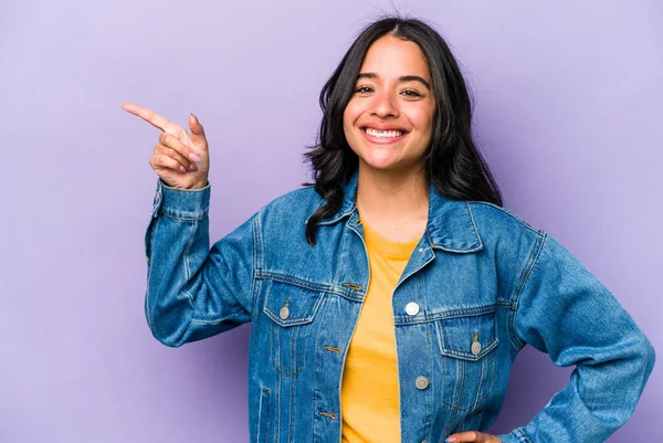 Mor arka planda izole edilmiş genç İspanyol kadın işaret parmağıyla işaret ederek gülümsüyor..