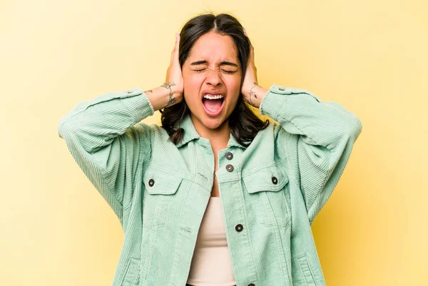 Sarı arka planda izole edilmiş genç İspanyol kadın kulaklarını elleriyle kapatıyor ve çok fazla ses duymamaya çalışıyor..