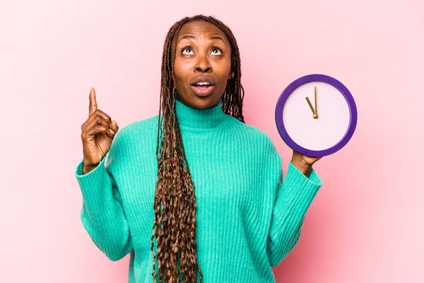 Genç Afrikalı Amerikalı bir kadın pembe arka planda izole edilmiş bir saat tutuyor ve ağzı açık bir şekilde üst tarafı işaret ediyor..