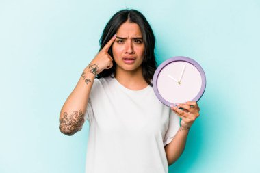 Mavi arka planda saati tutan genç İspanyol kadın işaret parmağıyla yaptığı hayal kırıklığı jestini gösteriyor..