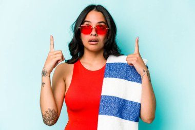 Elinde plaj havlusu tutan genç İspanyol kadın mavi arka planda izole edilmiş ağzı açık bir şekilde üst tarafı işaret ediyor..