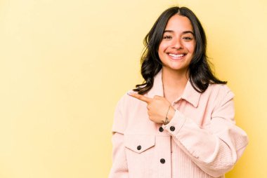 Sarı arka planda izole edilmiş genç İspanyol kadın gülümsüyor ve boş alanda bir şeyler gösteriyor..