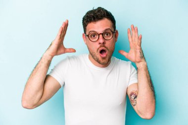 Mavi arka planda izole edilmiş genç beyaz bir adam zaferi ya da başarıyı kutluyor, şaşırıyor ve şok oluyor..