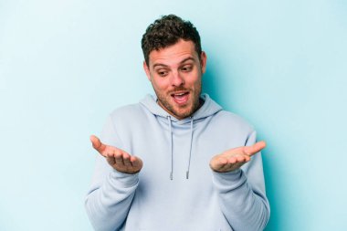 Kafkasyalı genç adam mavi arka planda izole edilmiş. Elinde avuç içi olan bir şey var ve kameraya sunuyor..