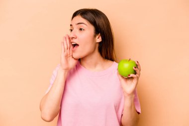 Elma tutan genç İspanyol kadın bej arka planda izole edilmiş bağırıyor ve avuç içini açık tutuyor..