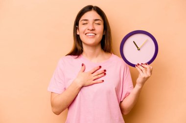 Latin kökenli genç bir kadın bej arka planda izole edilmiş bir saat tutuyor kahkahalar atıyor elini göğsüne koyuyor..