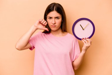 Latin kökenli genç bir kadın bej arka planda izole edilmiş bir saat tutuyor. Hoşlanmadığı bir jest gösteriyor. Anlaşmazlık konsepti.