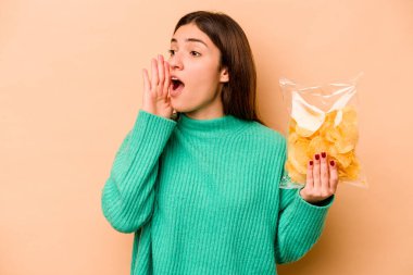 Latin kökenli genç bir kadın elinde bir çanta dolusu cipsle bej arka planda izole edilmiş bağırıyor ve ağzı açık bir şekilde avuç içi tutuyor..