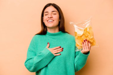 Bej arka planda izole edilmiş bir paket cipsi tutan genç İspanyol kadın kahkahalarla gülüyor..