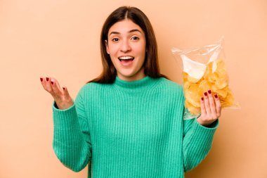 Latin kökenli genç bir kadın, elinde bej arka planda izole edilmiş bir paket cipsle hoş bir sürpriz bekliyor, heyecanlı ve el kaldırıyor..