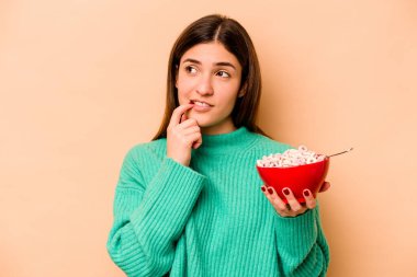 Latin kökenli genç bir kadın bej arka planda izole edilmiş mısır gevreği yiyor. Bir şeyin fotokopi odasına baktığını düşünüyor..