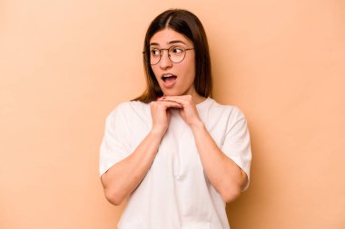 Latin kökenli genç bir kadın bej arka planda izole edilmiş şans için dua ediyor, şaşırmış ve ağzını açıyor..