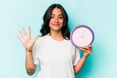 Mavi arka planda bir saat tutan genç İspanyol kadın gülümseyerek beş numarayı parmaklarıyla gösteriyor..