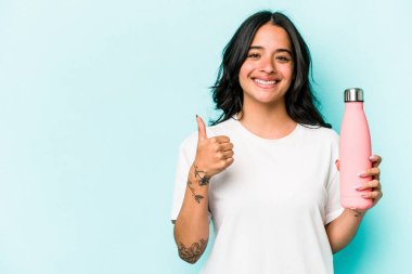 Mavi arka planda termo tutan genç İspanyol kadın gülümsüyor ve başparmağını kaldırıyor.