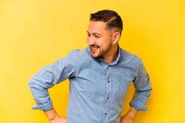 Sarı arka planda izole edilmiş genç İspanyol adam mutlu bir şekilde gülüyor ve ellerini karnının üstünde tutarak eğleniyor..