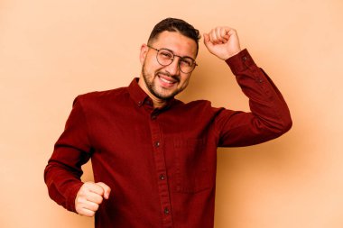 Latin kökenli genç adam, bej arka planda izole edilmiş özel bir günü kutluyor, atlıyor ve enerjiyle kollarını kaldırıyor..