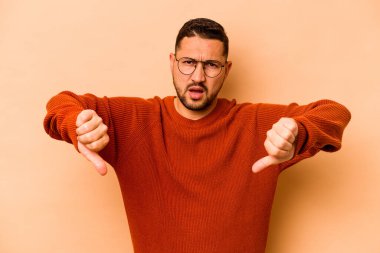 Latin kökenli genç adam bej arka planda izole edilmiş baş parmağını aşağı gösteriyor ve hoşlanmadığını ifade ediyor..