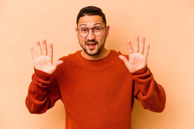 Latin kökenli genç bir adam bej arka planda izole edilmiş. Hoş bir sürpriz alıyor, heyecanlı ve el kaldırıyor..