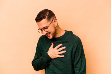 Latin kökenli genç bir adam bej arka planda izole edilmiş. Gülüyor, elini kalbe koyuyor, mutluluk kavramını koruyor..