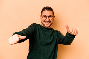 Latin kökenli genç adam bej arka planda izole edilmiş. Baş parmağını kaldırıyor, gülümsüyor ve kendinden emin..