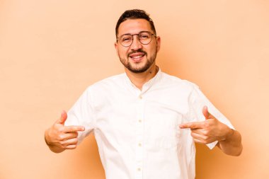 Latin kökenli genç bir adam bej arka planda izole edilmiş. Elle bir tişört fotokopi alanını işaret ediyor, gururlu ve kendinden emin.