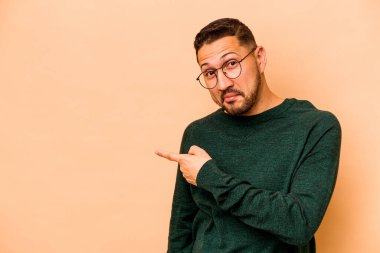 Latin kökenli genç adam bej arka planda izole edilmiş gülümsüyor ve boş alanda bir şeyler gösteriyor..