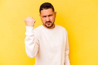 Sarı arka planda izole edilmiş genç İspanyol adam kameraya yumruk gösteriyor, agresif yüz ifadesi..