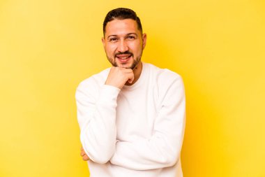 Sarı arka planda izole olmuş genç İspanyol adam gülümsüyor ve kendinden emin, elleriyle çeneye dokunuyor..