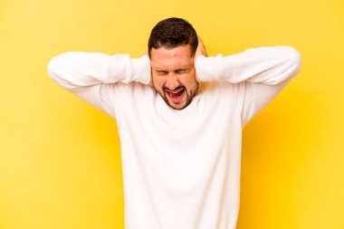 Latin kökenli genç adam sarı arka planda izole edilmiş kulakları kulaklarıyla kapatıyor çok fazla ses duymamaya çalışıyor..