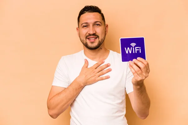 Hispanic man holding wifi placard isolated on beige background laughs out loudly keeping hand on chest.