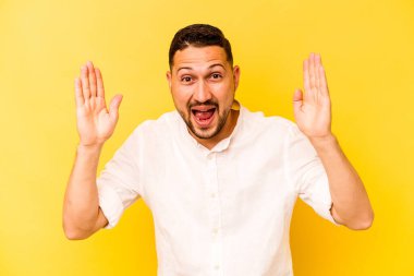 Latin kökenli genç adam sarı arka planda izole edilmiş. Hoş bir sürpriz bekliyor. Heyecanlı ve el kaldırıyor..