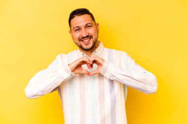 Sarı arka planda izole edilmiş genç İspanyol adam gülümsüyor ve elleriyle kalp şeklini gösteriyor..