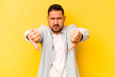 Sarı arka planda izole edilmiş genç İspanyol adam baş parmağını aşağı gösteriyor ve hoşlanmadığını ifade ediyor..