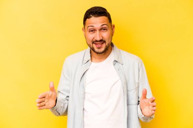 Sarı arka planda izole edilmiş genç İspanyol adam iki eliyle bir şey tutuyor, ürün sunumu..