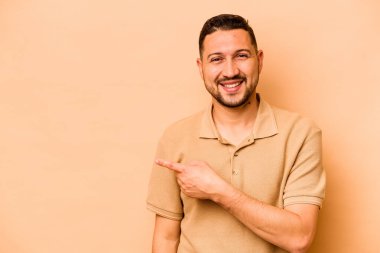Latin kökenli genç adam bej arka planda izole edilmiş gülümsüyor ve boş alanda bir şeyler gösteriyor..