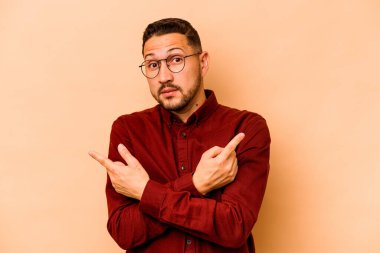 Latin kökenli genç adam bej arka planda iki seçenek arasında seçim yapmaya çalışıyor..