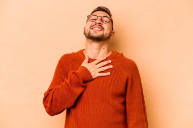 Latin kökenli genç adam bej arka planda izole edilmiş. Yüksek sesle gülüyor. Elini göğsünde tutuyor..