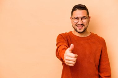 Genç İspanyol adam bej arka planda izole edilmiş gülümsüyor ve başparmağını kaldırıyor.