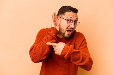 Latin kökenli genç bir adam bej arka planda bir dedikoduyu dinlemeye çalışıyor..