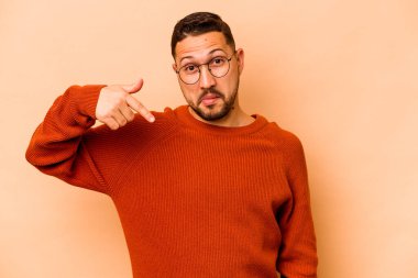 Latin kökenli genç bir adam bej arka planda izole edilmiş. Elle bir tişört fotokopi alanını işaret ediyor, gururlu ve kendinden emin.