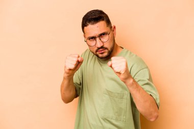 Latin kökenli genç adam bej arka planda izole edilmiş. Kameraya yumruk, agresif yüz ifadesi gösteriyor..