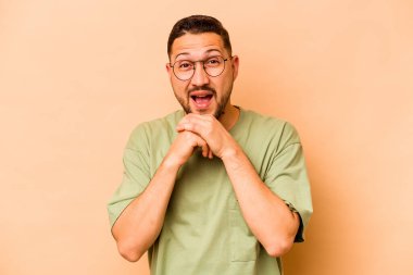 Latin kökenli genç adam bej arka planda izole edilmiş şans için dua ediyor, şaşırmış ve ağzını açıyor..