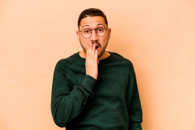 Latin kökenli genç bir adam, bej arka planda izole edilmiş şok olmuş, ağzı ellerle kaplanmış, yeni bir şey keşfetmeye hevesli..